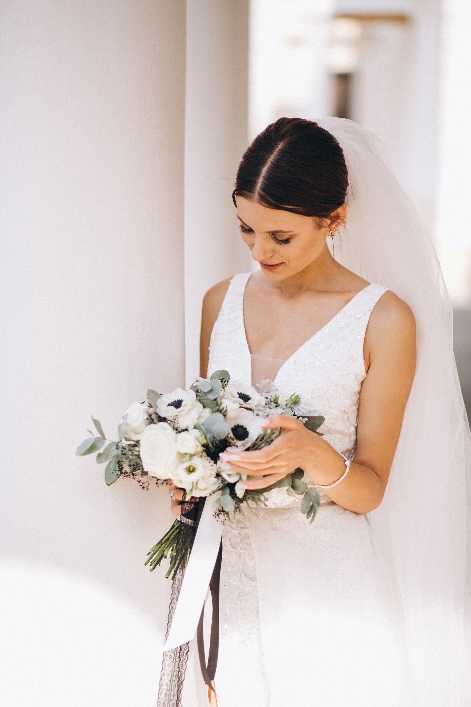 Bride in white dress holds elegant bouquet of white flowers and eucalyptus against light background.