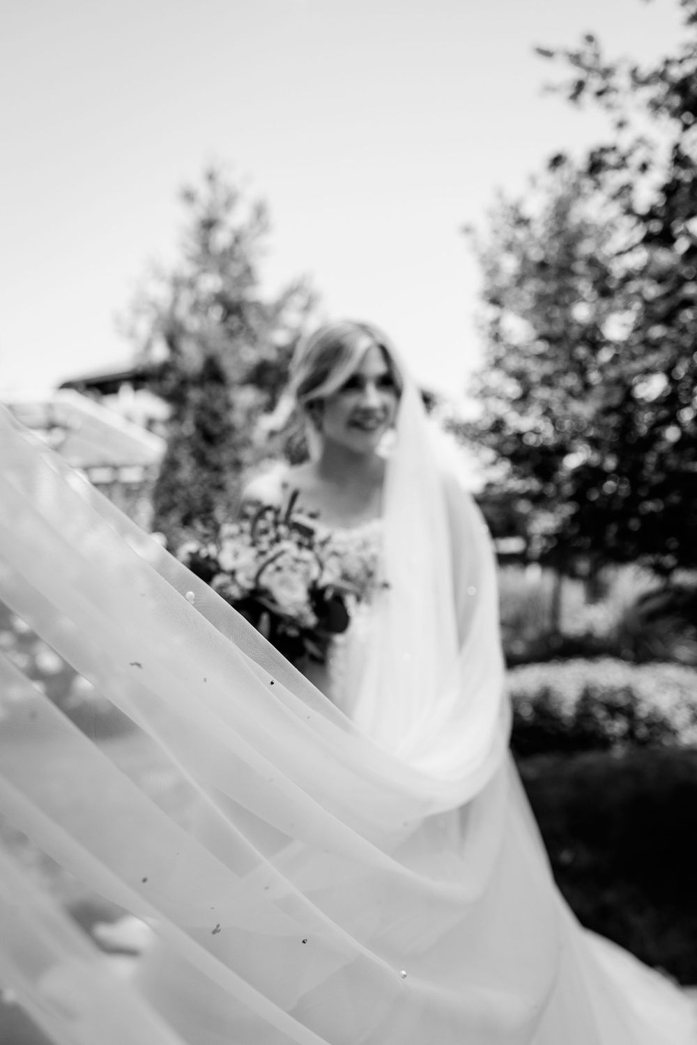 Artistic black and white shot of bride with flowing veil in outdoor garden.