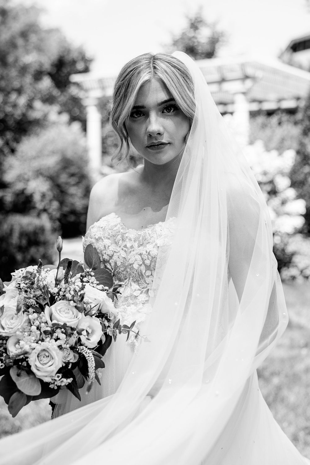 Black and white bridal portrait with delicate veil and floral bouquet in garden setting.