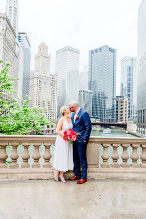 married couple in navy and silver with red bouquet kissing in front of Chicago River