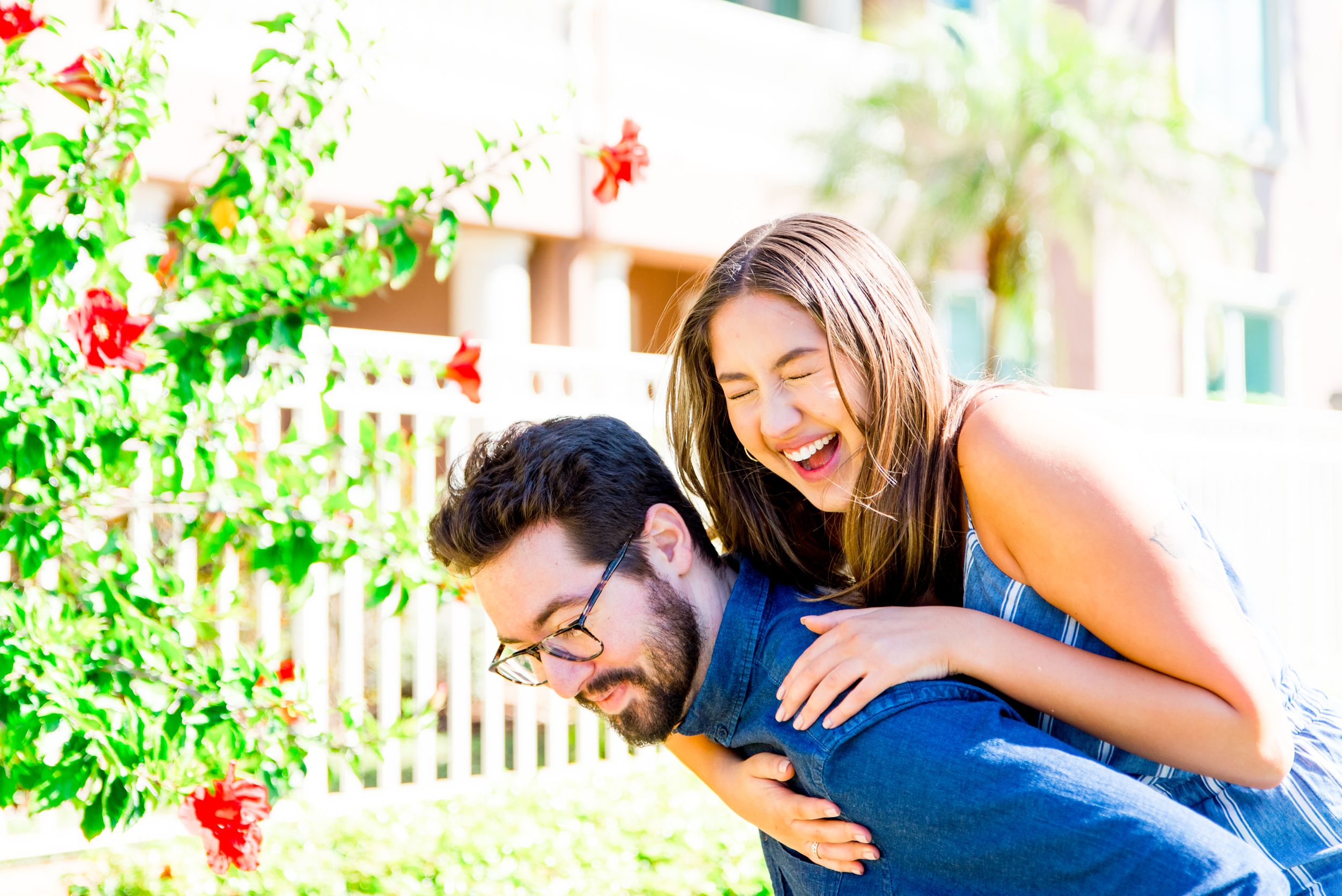 young woman piggyback on boyfriend, laughing next to pink hibiscus tree at Vinoy Park sunrise engagement pictures