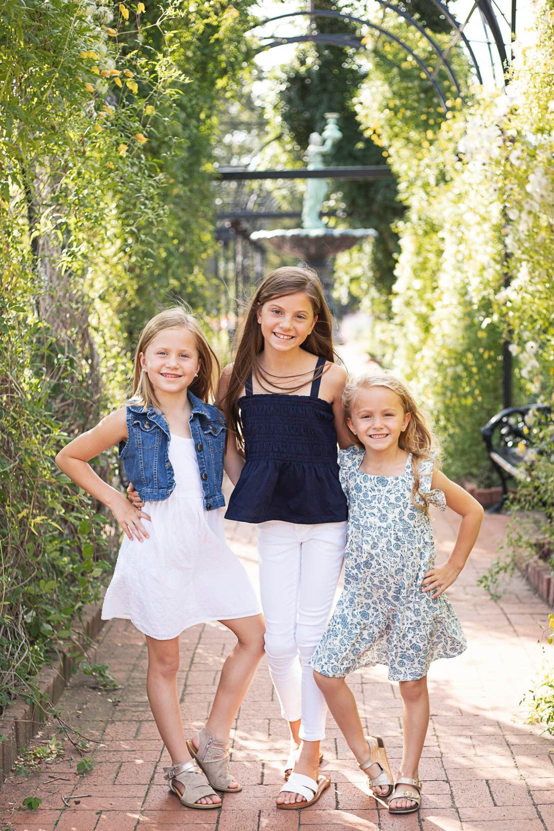 three sisters smiling and posing for the camera. standing in a greenery arch in the summer. Wearing blue and white.