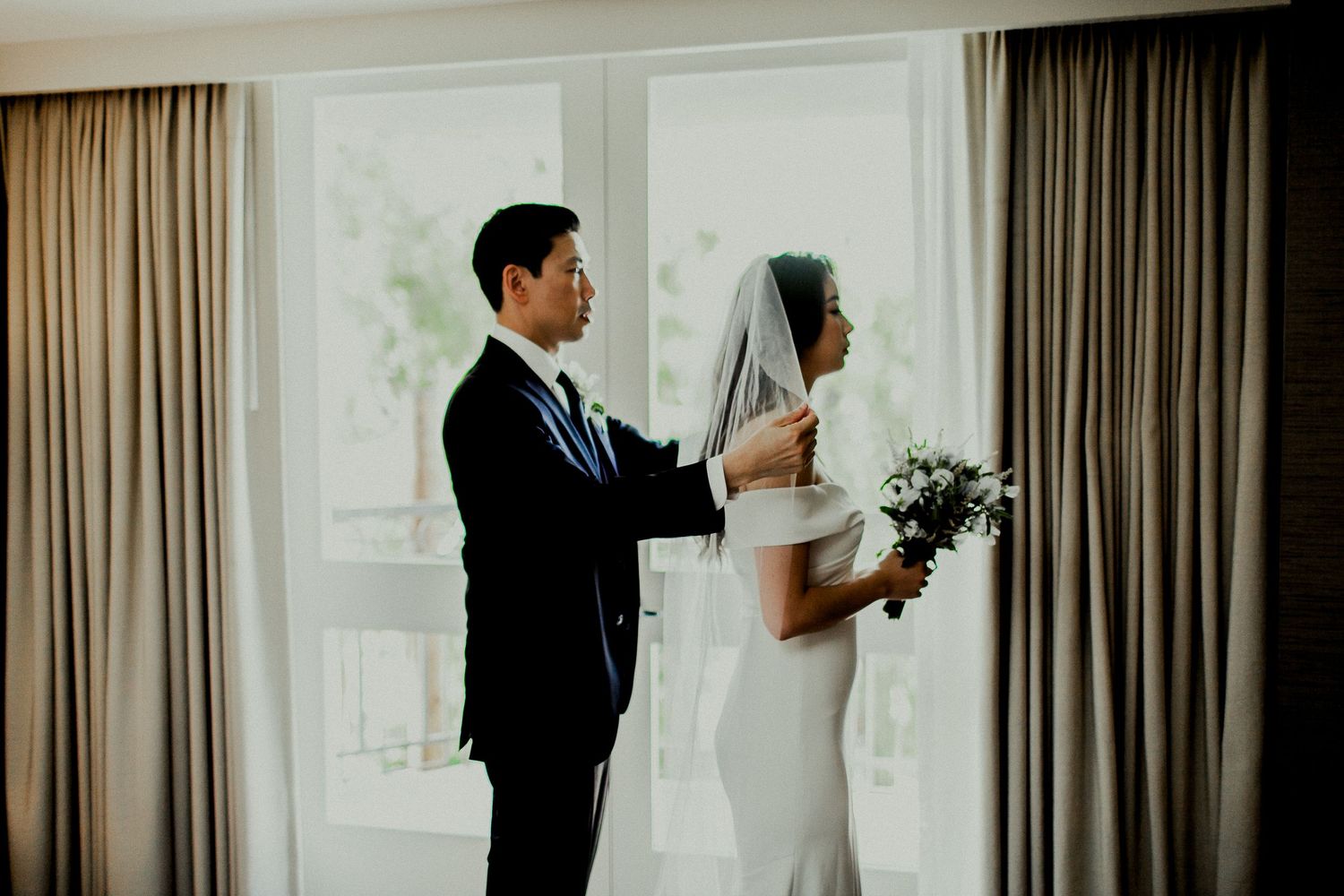 groom adjusting veil for bride in front of balcony window