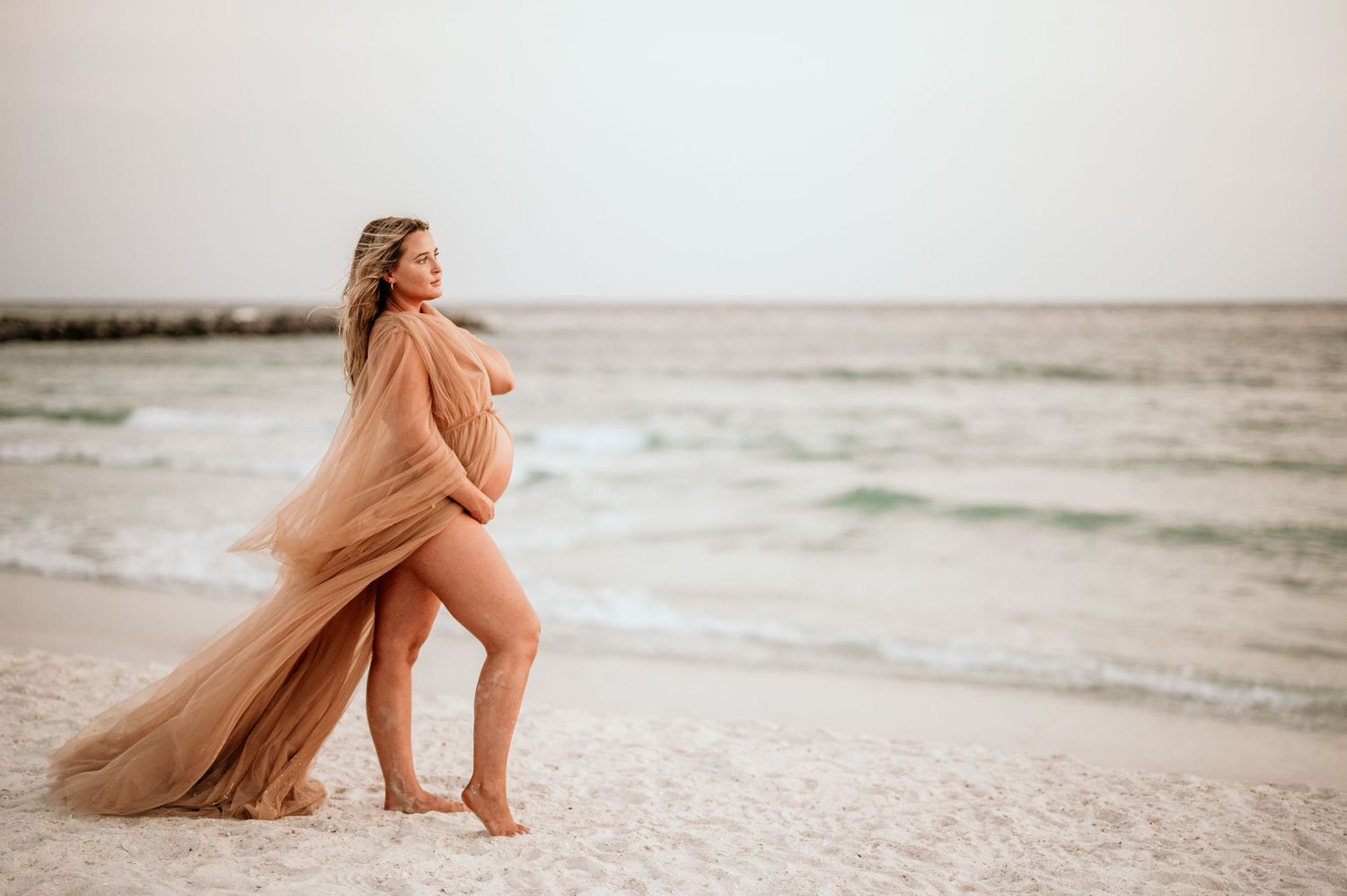 Woman in flowing peach dress poses elegantly on beach at sunset.