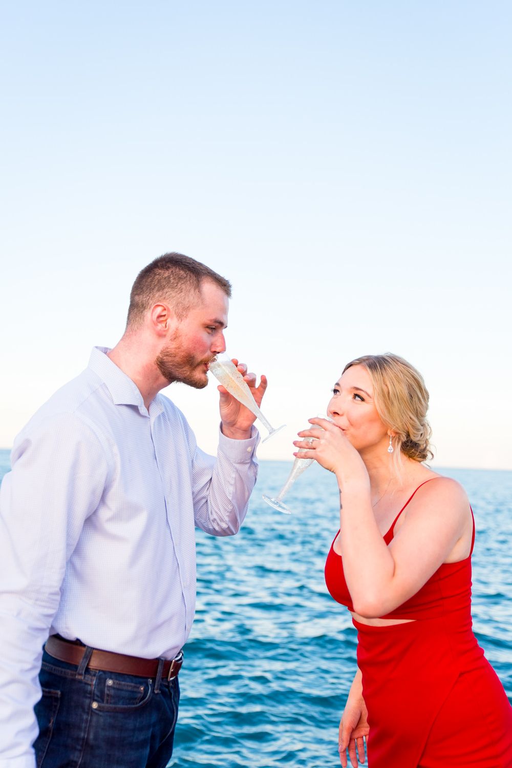 woman in red dress and man in white shirt sip champagne at Chicago sunset engagement pictures by the lakefront trail