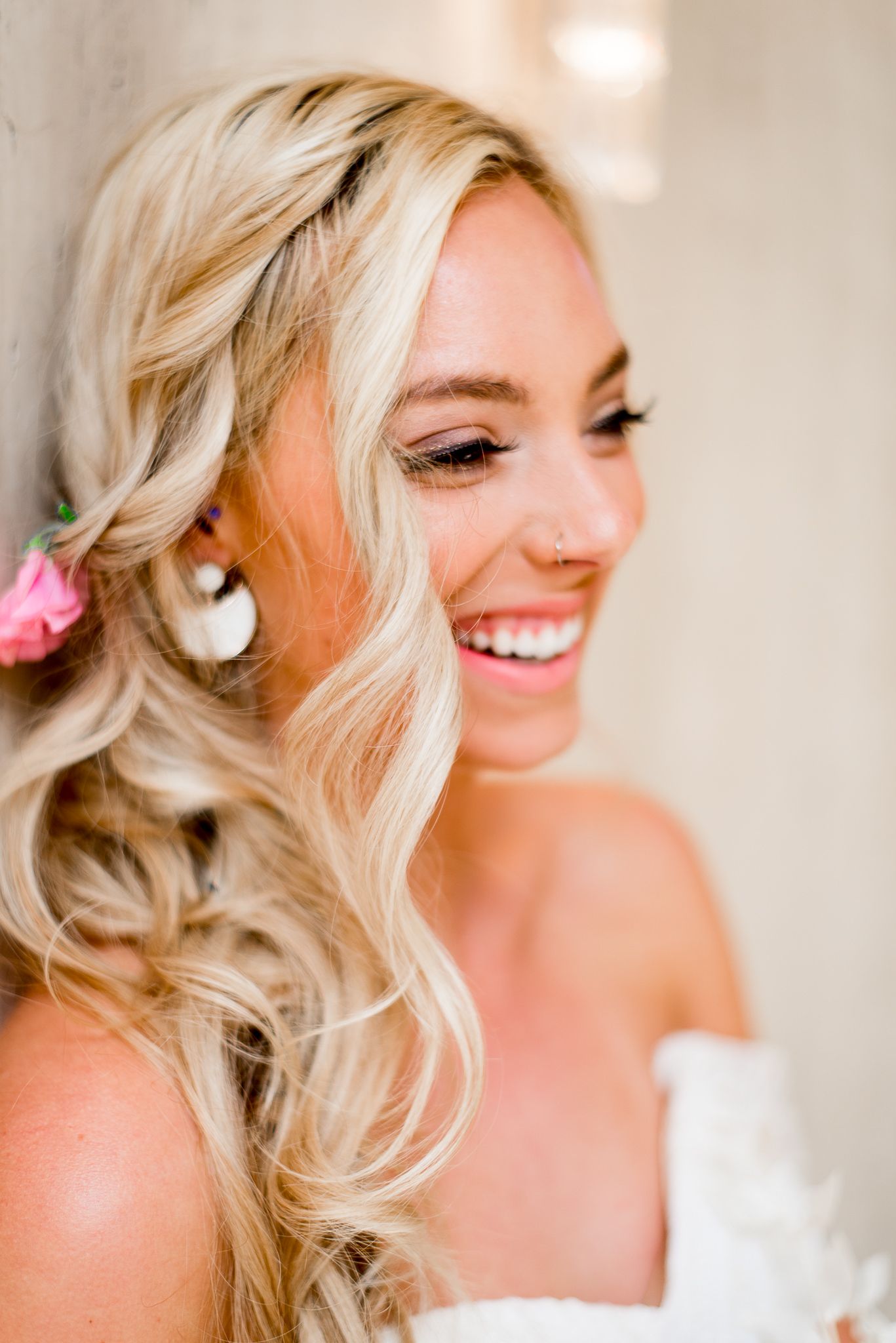 blonde bride with braided hair, pink flower in hair, white earrings, and glitter eyeliner smiling and giggling