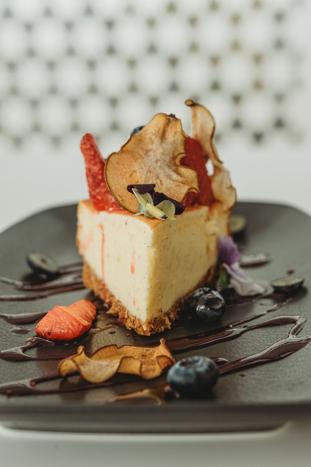 Elegant cheesecake slice topped with berries, and chocolate drizzle on a dark plate. SIX84 Photography, Food Photography, Sydney