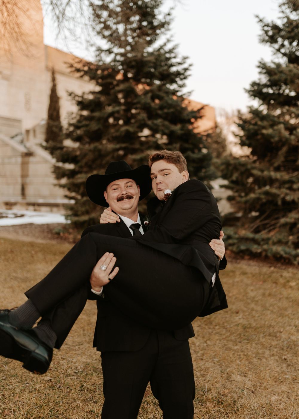 Playful formal portrait of two people in black suits against evergreen trees at sunset.