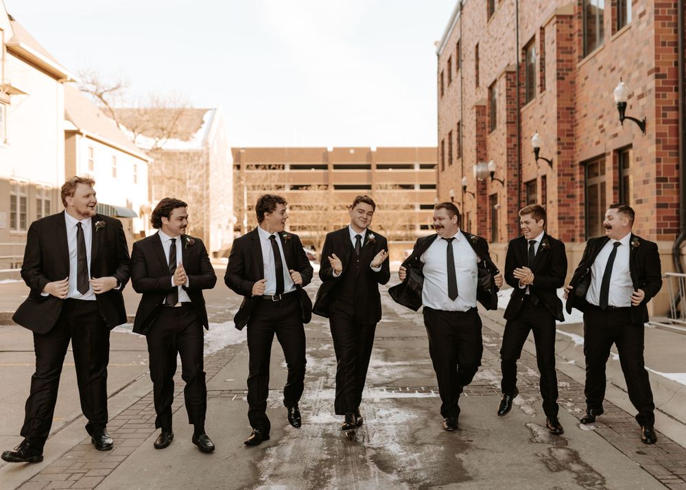Groomsmen in black suits and ties pose playfully in an urban setting.