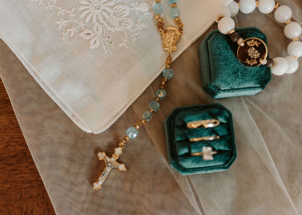 Wedding accessories including embroidered handkerchief, rosary, and teal ring boxes on wooden surface.