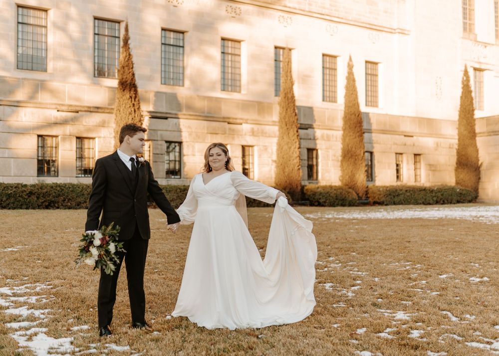 Wedding couple poses playfully on snowy grounds in front of a stately building.