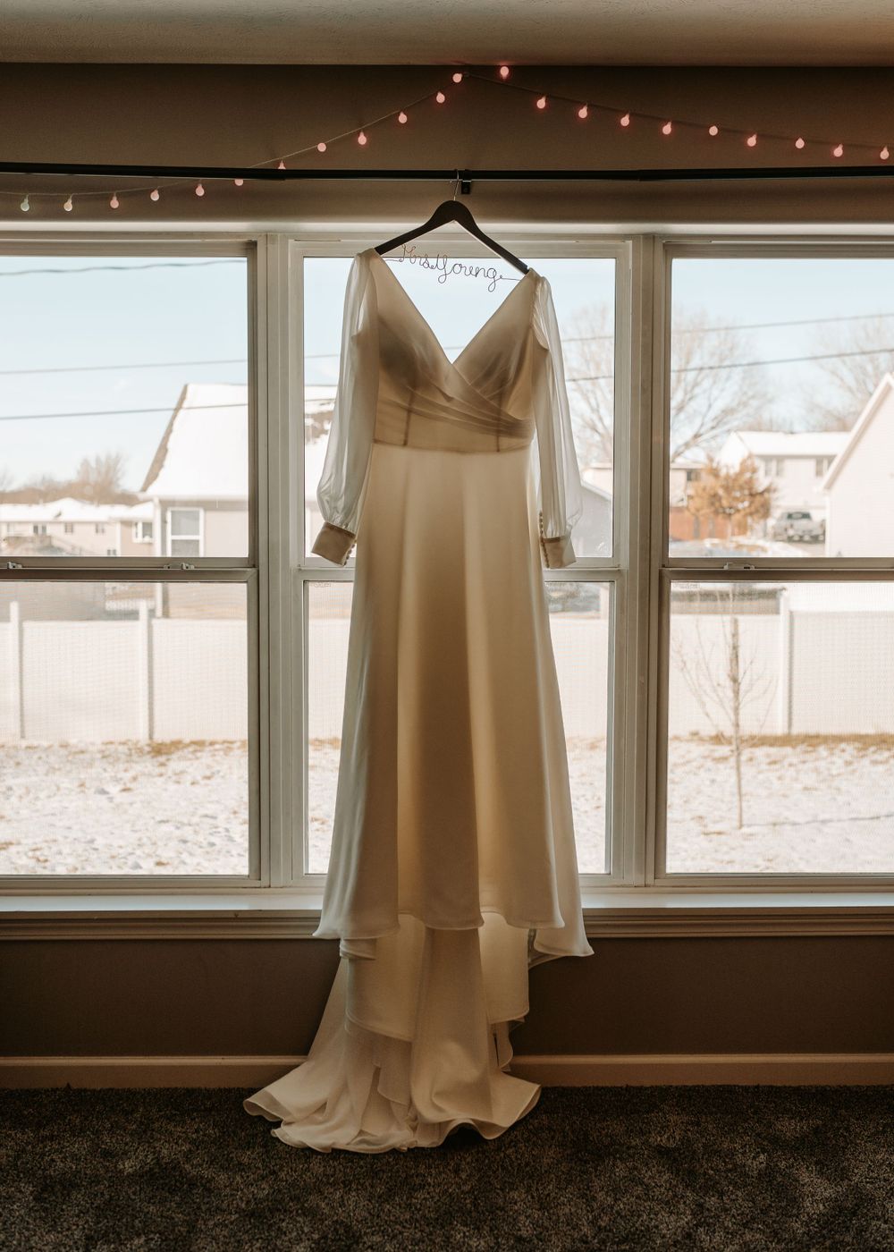 White wedding dress hanging in window with string lights above and winter scene outside.