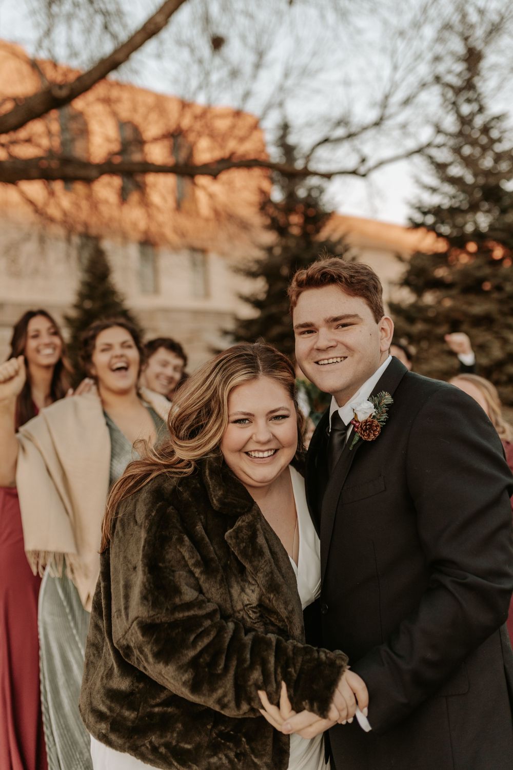 Winter wedding couple in formal attire embracing outdoors with evergreen trees in background.