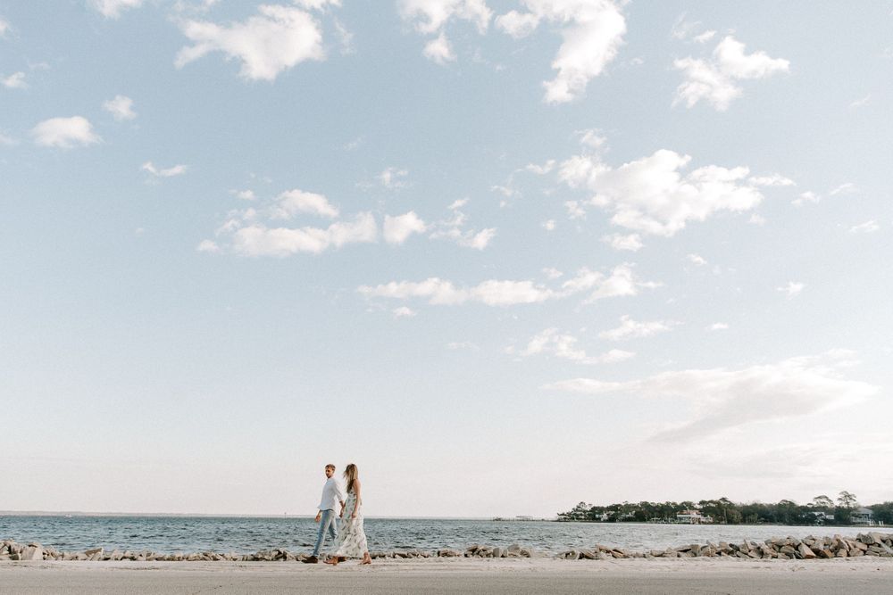 couple holding hands and waling in front of the water during their sunrise engagement photoshoot