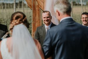groom smiling at bride shot between the backs of bride and father of the bride