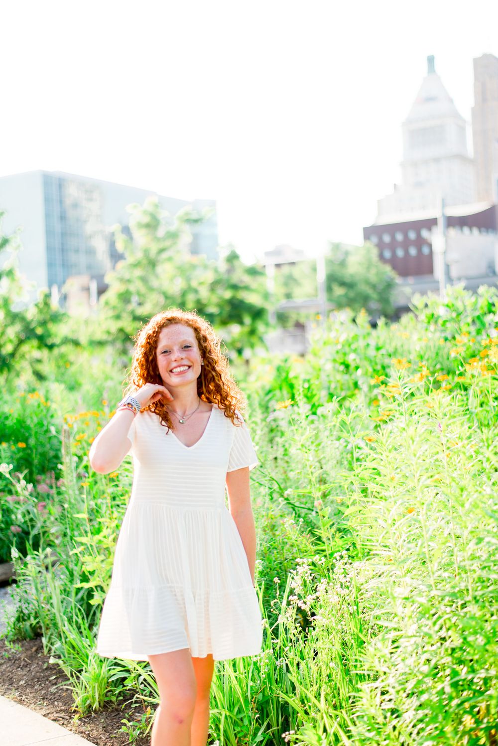 young woman in white dress flips her hair back in front of flowers and cityscape at Smale Park senior pictures