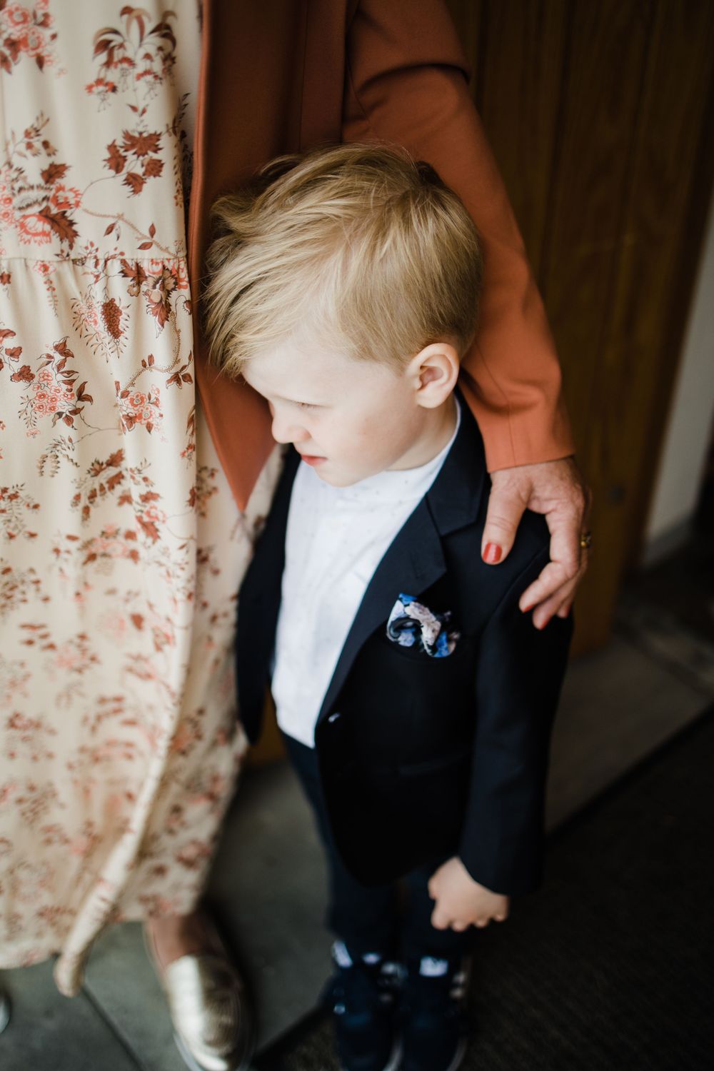 A young boy in formal attire stands against a floral curtain during a wedding.