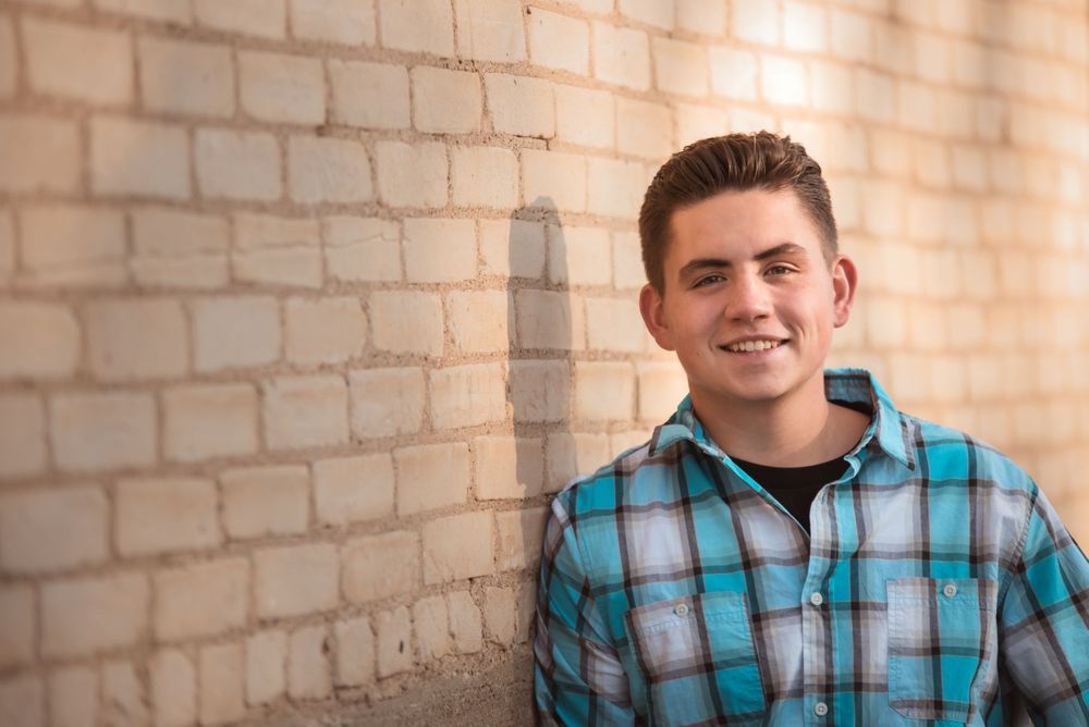 Boy with black hair wearing turquoise white black plaid shirt leaning against yellow brick wall