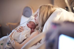 A mother with long blonde hair sits with eyes closed in a hospital bed holding her newborn skin to skin on her chest.