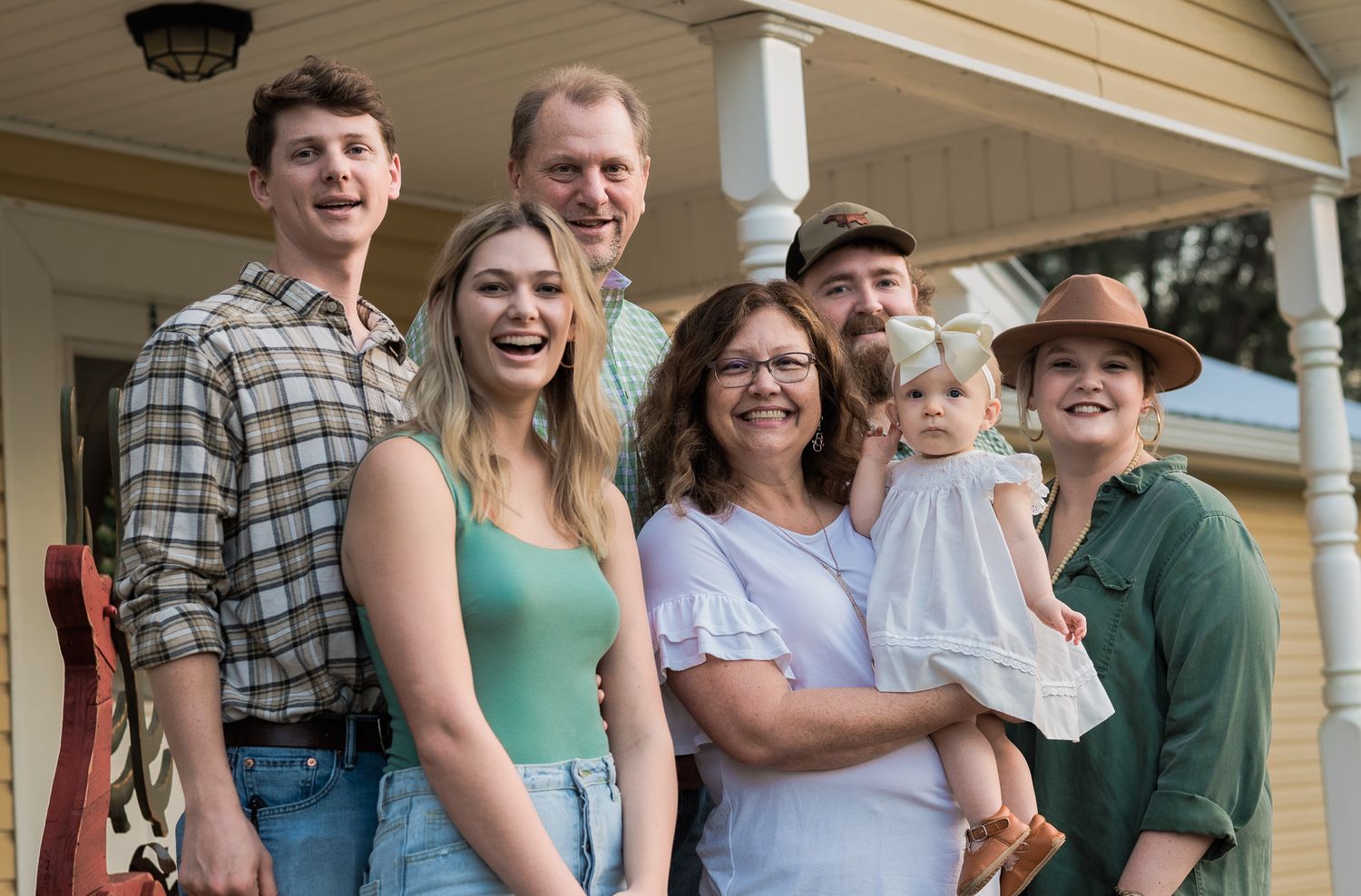 Multigenerational family group stands together on a covered porch outside a house.