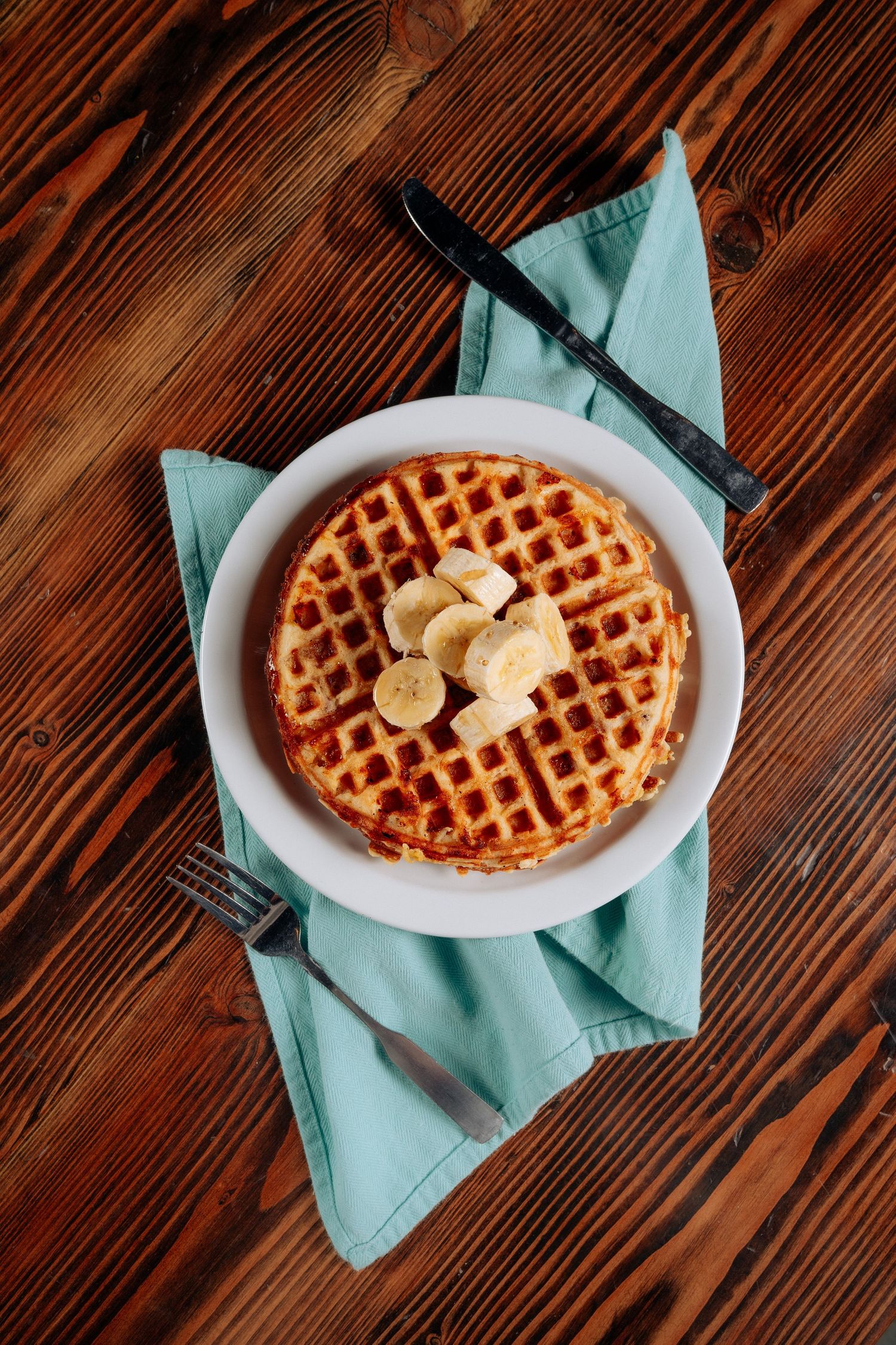 Golden waffle topped with butter and bananas on turquoise napkin against wooden surface.