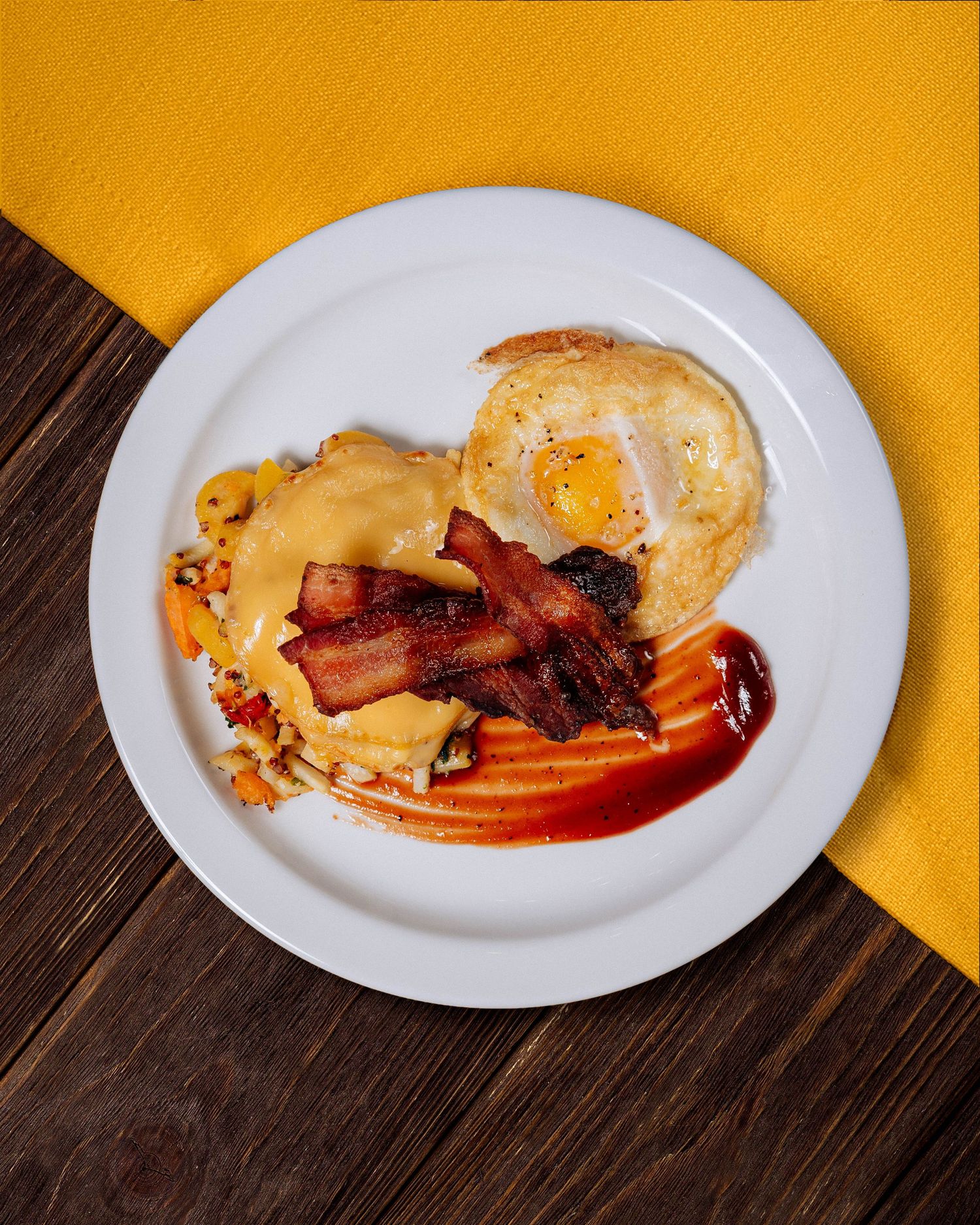 Eggs Benedict with bacon and hollandaise sauce on white plate against yellow background.