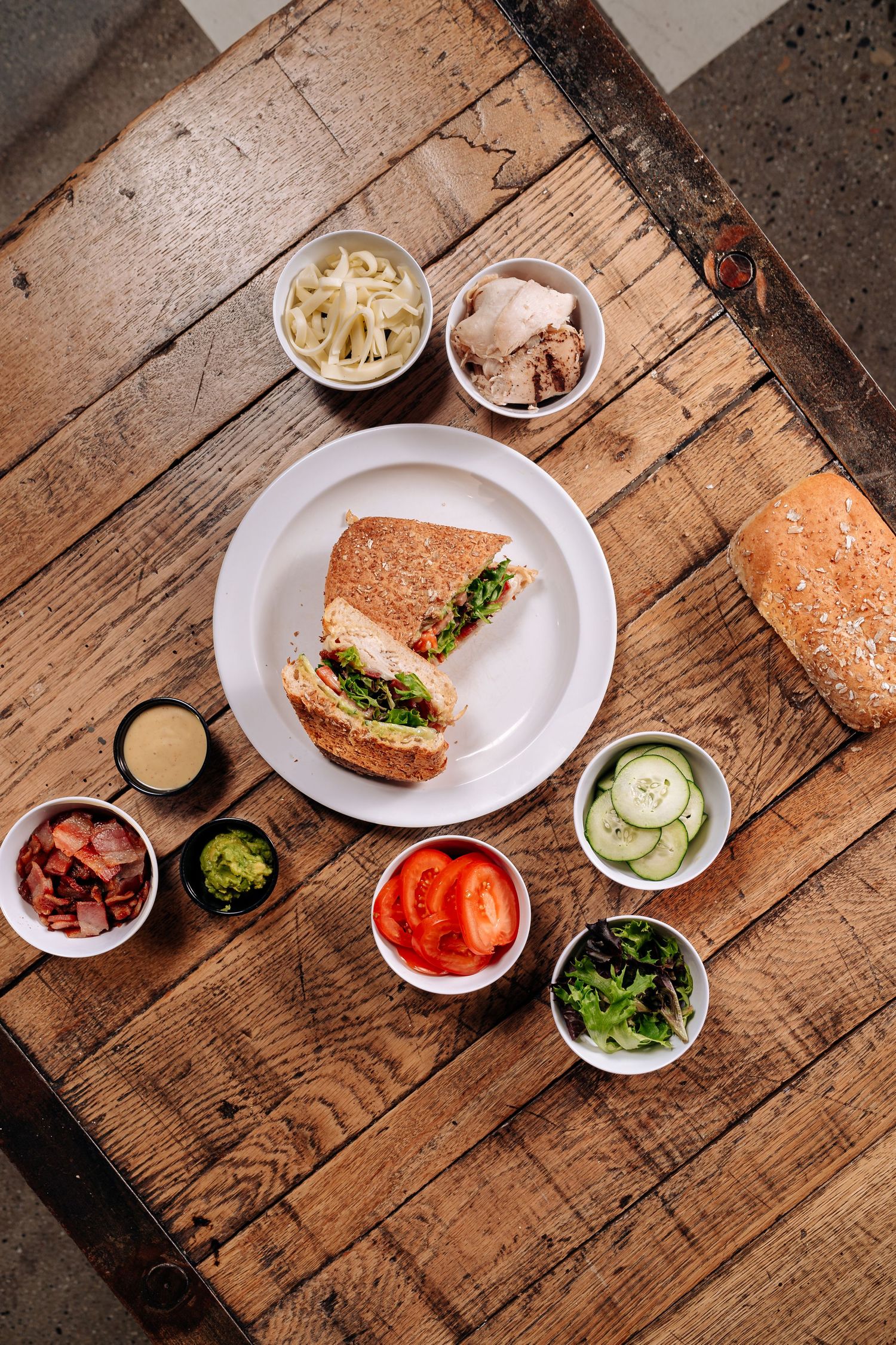 Sandwich on white plate surrounded by assorted condiments and toppings on wooden surface.