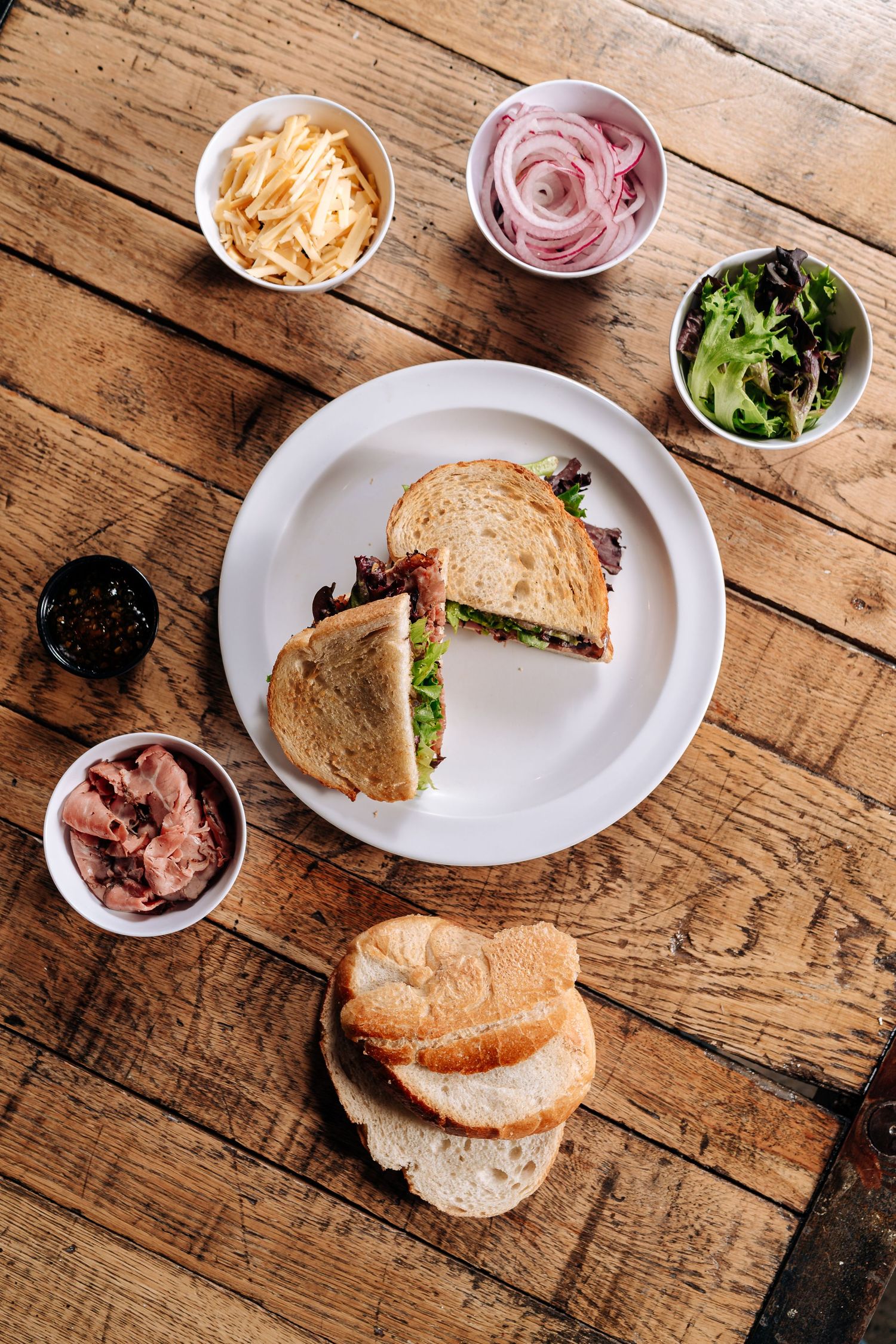 Halved sandwich on plate with various toppings and condiments in small bowls on wooden table.