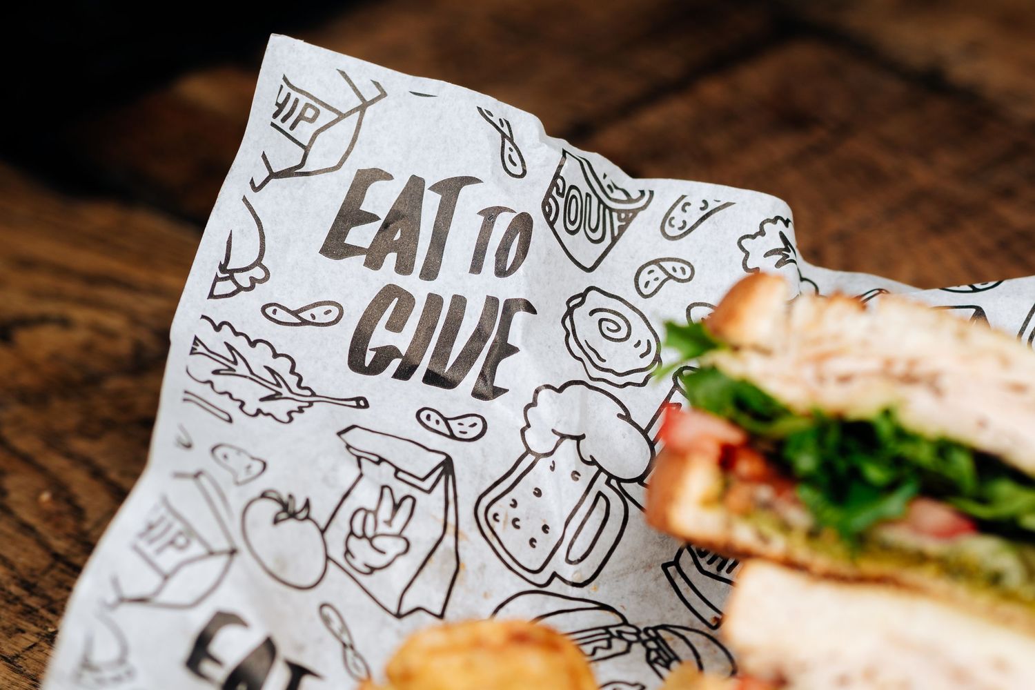 Restaurant wrapper with 'Eat To Give' text and food doodles next to sandwich.