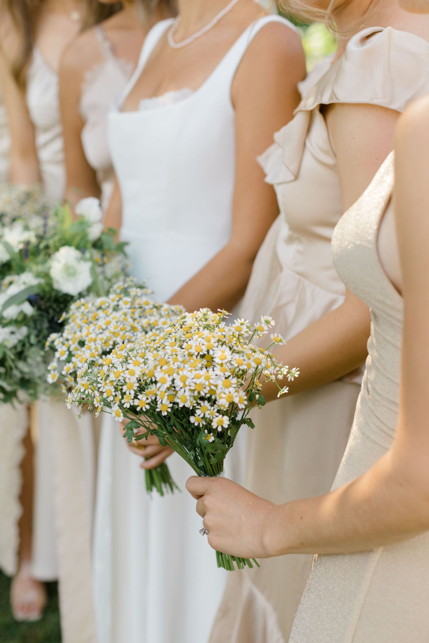 Bridesmaids in cream dresses holding fresh daisy bouquets at an outdoor wedding ceremony.