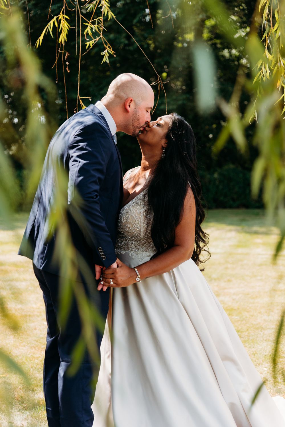 A romantic kiss captured through hanging willow branches on a sunny day.