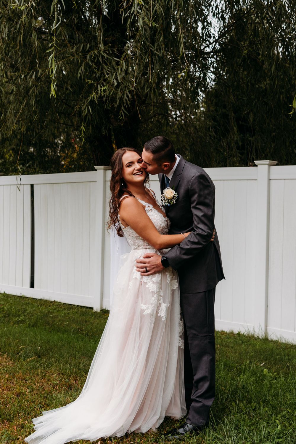A romantic wedding moment captured against a white fence and willow tree.