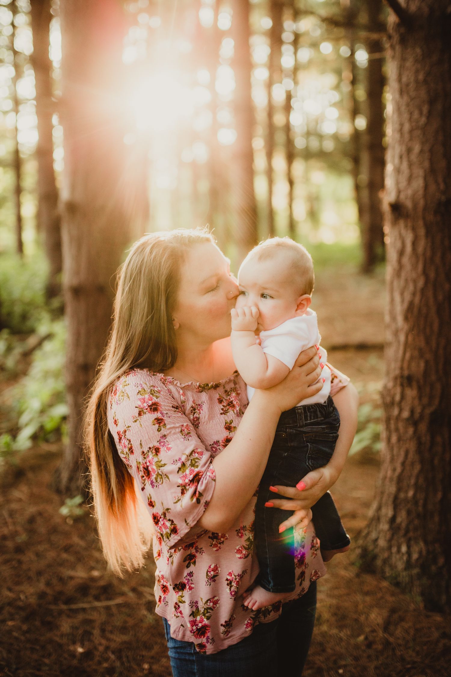 a mother stands in the forest at golden hour; her hair glows in the sunlight as she holds her baby and kisses his cheek