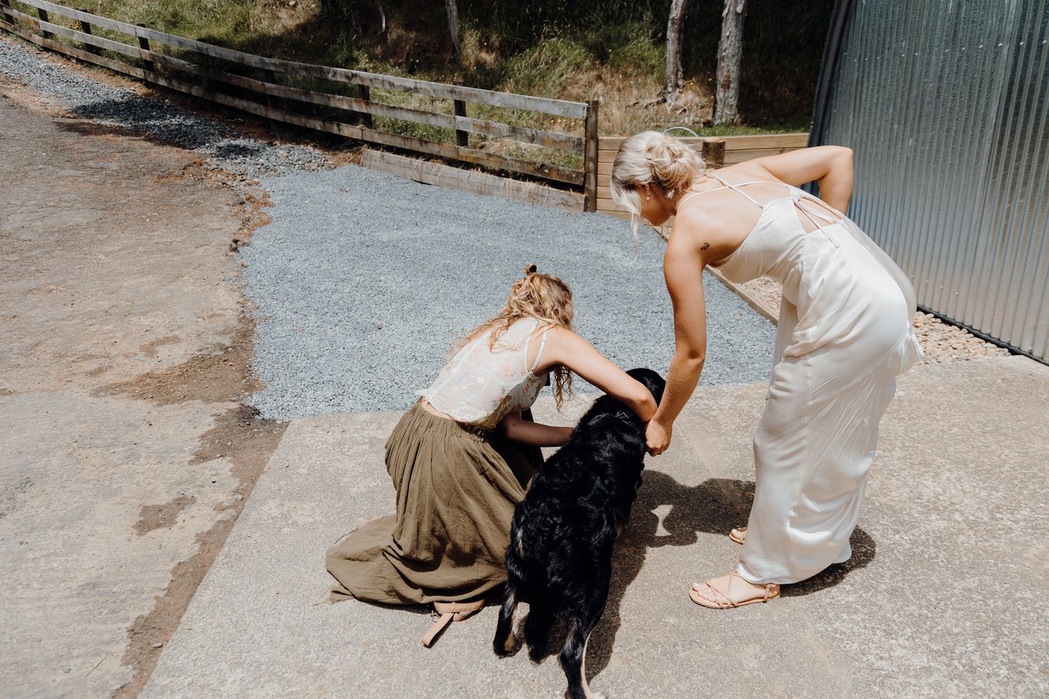 People petting a black dog on a concrete path outside a rural building.
