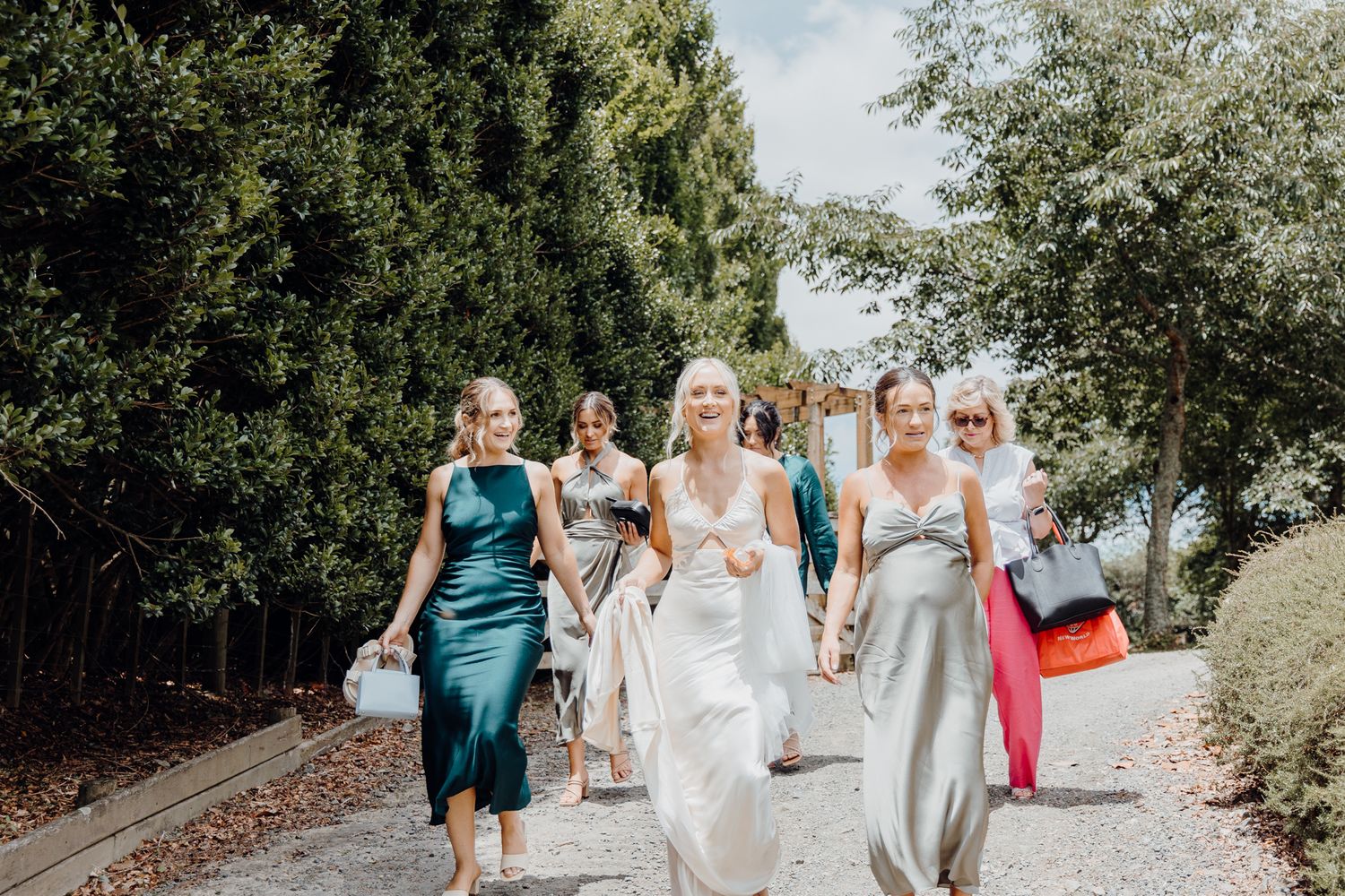 Wedding party walking along a sunny outdoor path carrying colorful bags and bouquets.