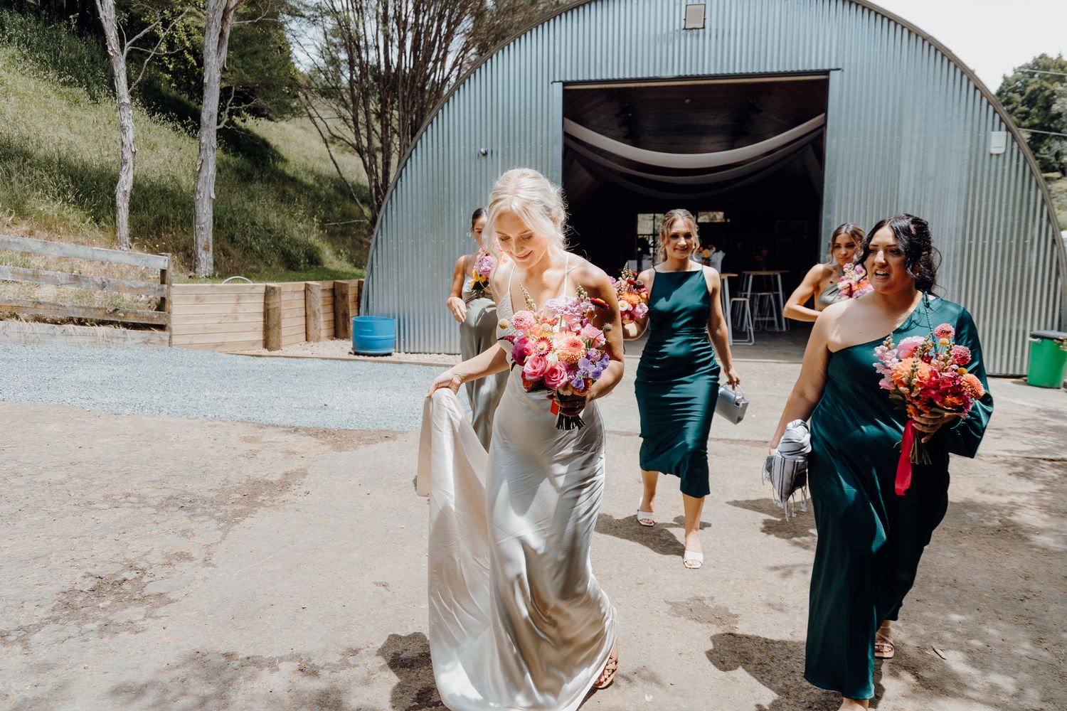 Wedding party emerging from a blue quonset hut carrying vibrant floral bouquets.