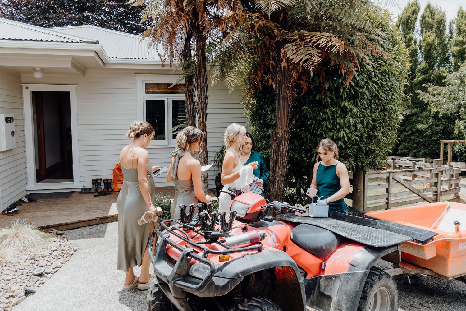 Wedding party gathered around an orange ATV outside a white house on a sunny day.