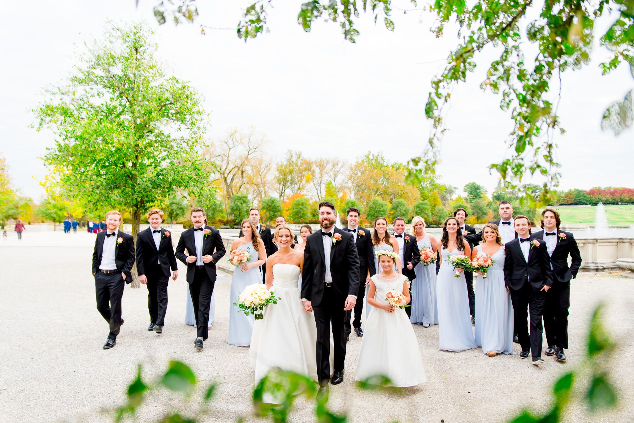 large wedding party in black suits and light blue dresses walk with bride and groom at Forest Park STL fall wedding