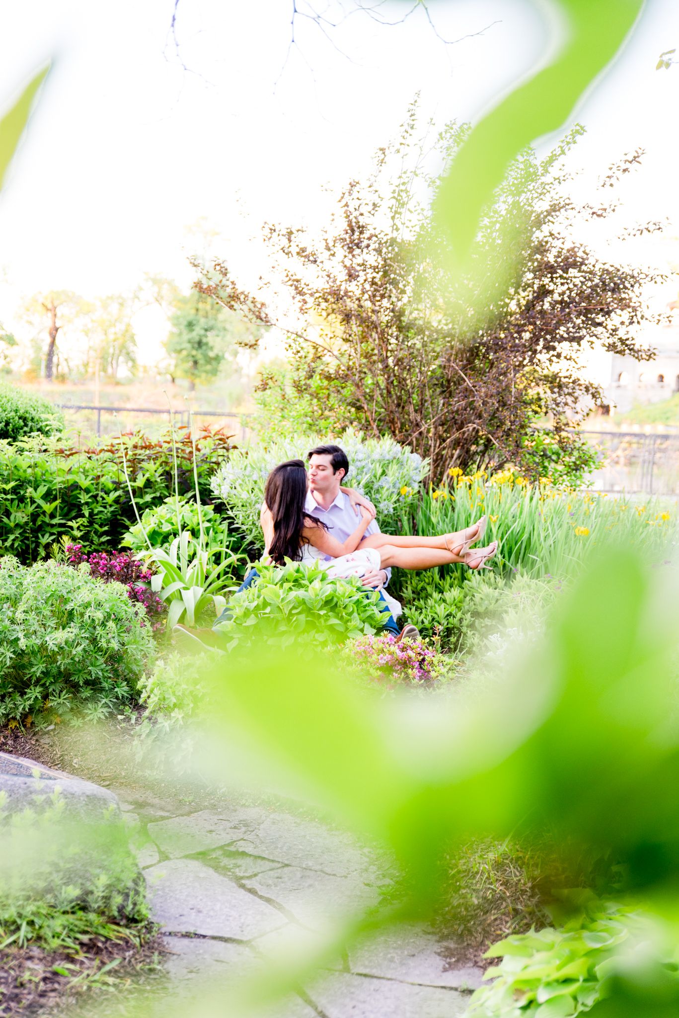 woman sitting on fiance's lap in Lincoln Park as they kiss for engagement pictures in the garden
