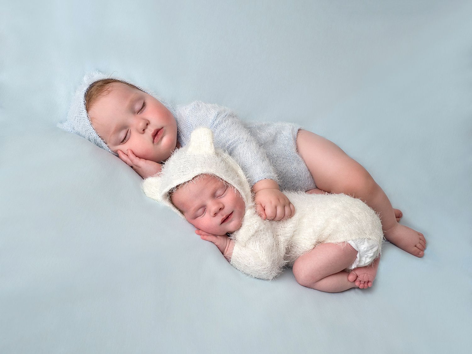 A one year old and newborn baby lay sleeping together dressed in teddy bear suits.