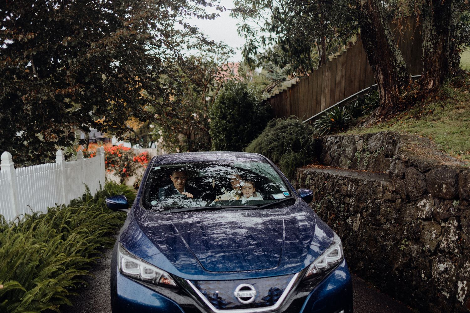 A blue Nissan sedan parked on a residential street with stone walls and ferns.