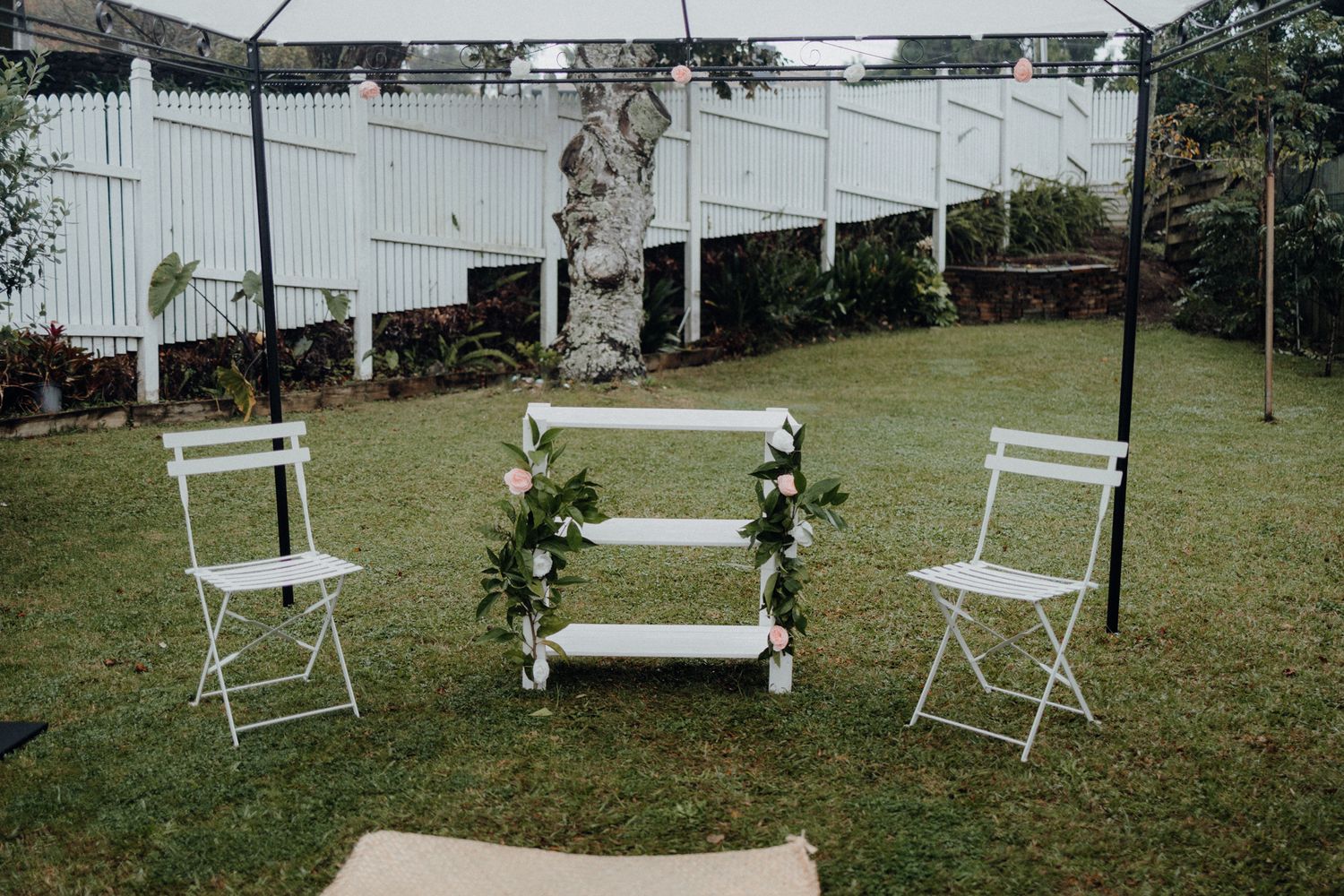 Simple outdoor wedding ceremony setup with white chairs and floral bench in garden setting.