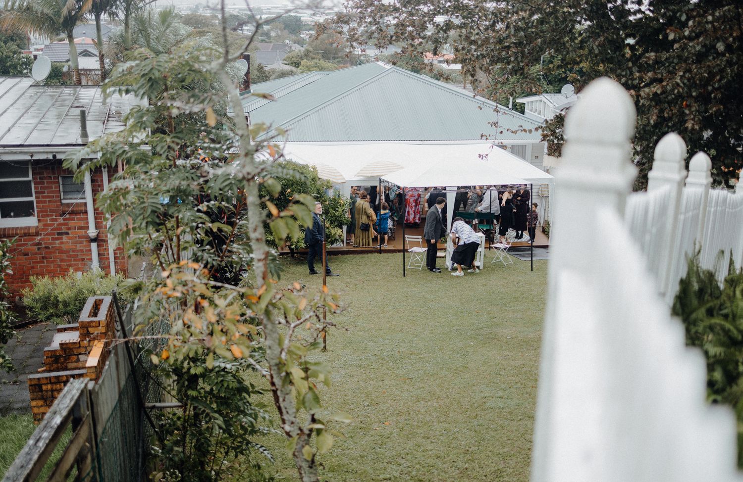 Backyard wedding reception setup with white tent and picket fence in residential setting.