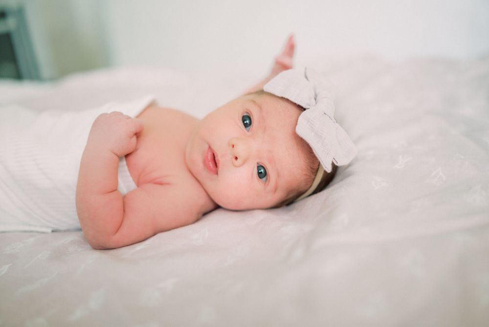 Baby wearing pink bow headband lying on white bedding during photoshoot.