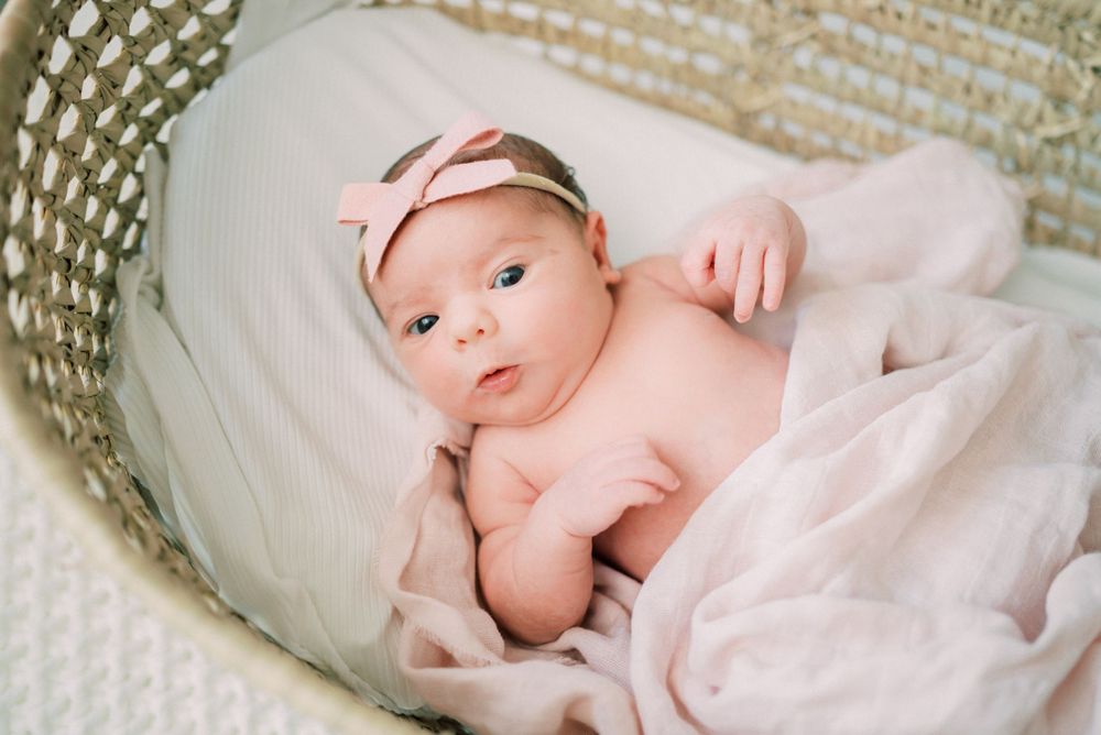 Baby wearing pink bow headband lying in woven basket on white blanket.