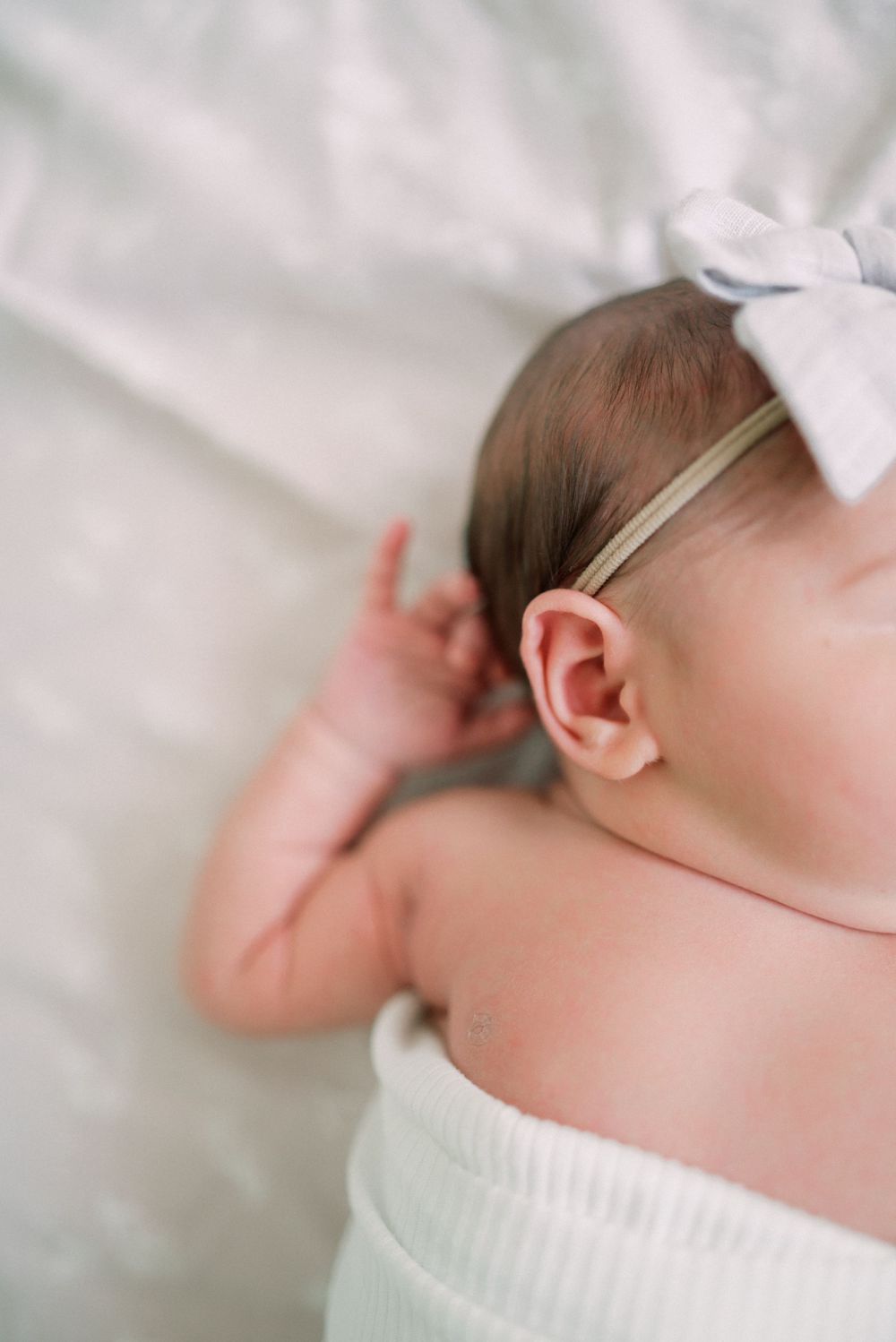 Close-up detail of baby's ear with white bow headband against white fabric.