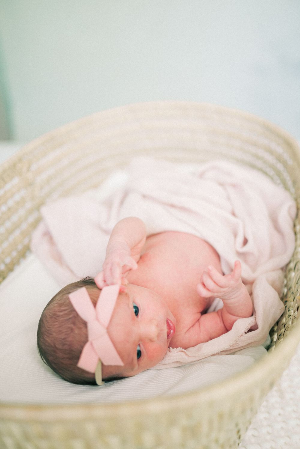 Newborn baby with pink bow lying in woven basket wrapped in light fabric.