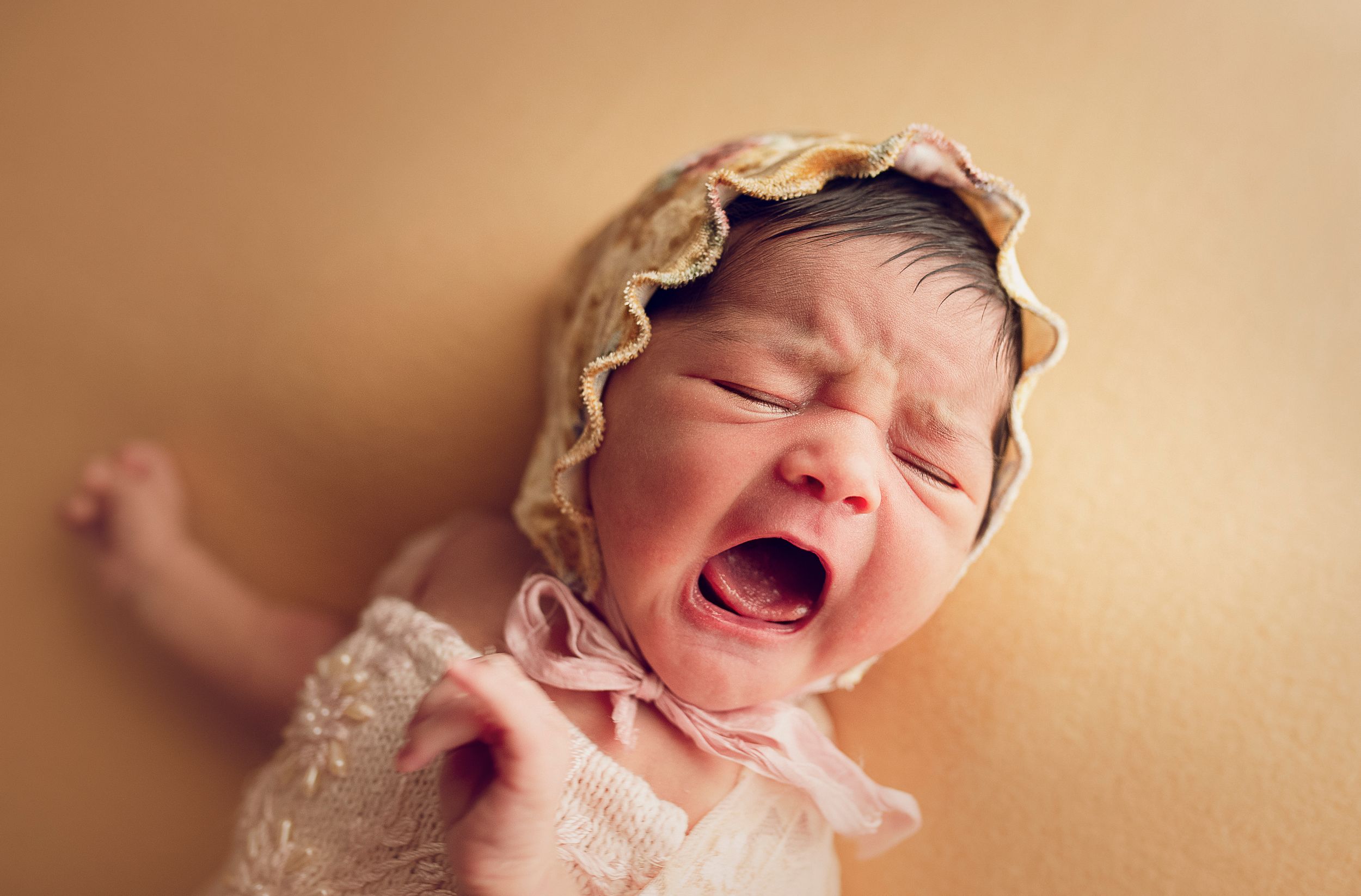 newborn baby wearing a bonnet crying peach backdrop