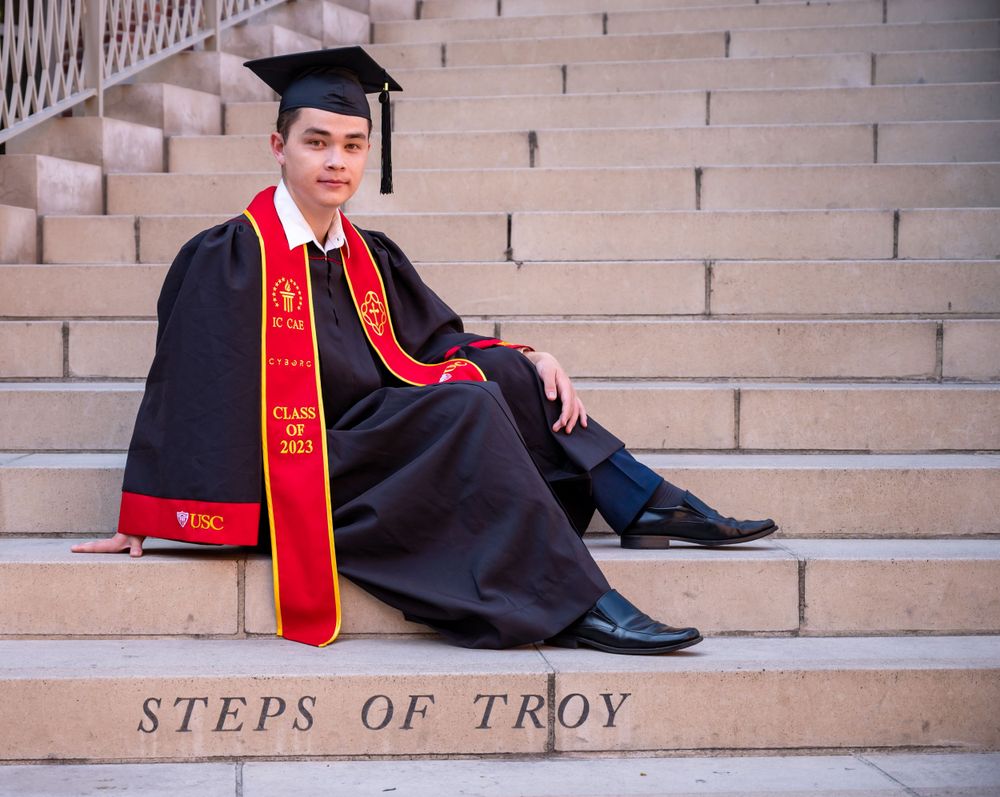Graduate in cap and gown sitting on concrete steps labeled 'Steps of Troy'.