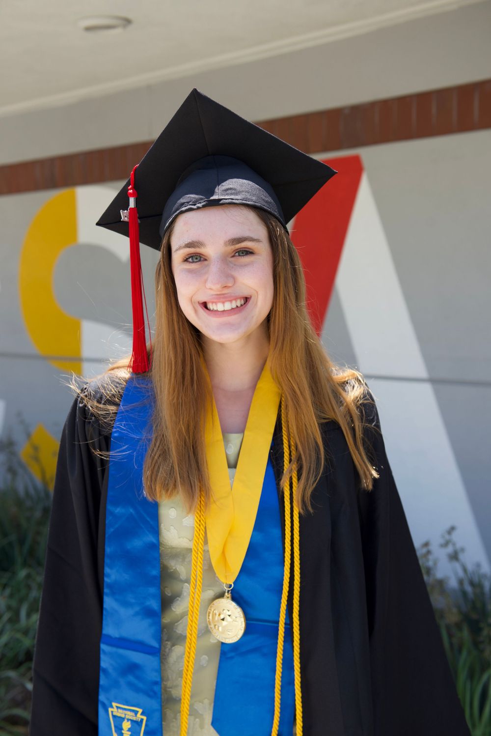 Graduate wearing cap, gown and medal stands in front of colorful wall.